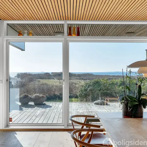 En lys stue med moderne møbler, hvor farverige glasfigurer står ved en gul sofa. Store vinduer giver udsigt over havet. Bøger fylder en reol ved siden af fjernsynet.