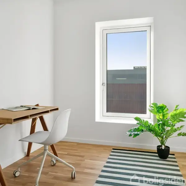 Et skrivebord med åben bog står ved siden af en hvid stol i et lyst rum. Et vindue til højre tillader naturligt lys. En grøn plante og stribet tæppe dekorerer.