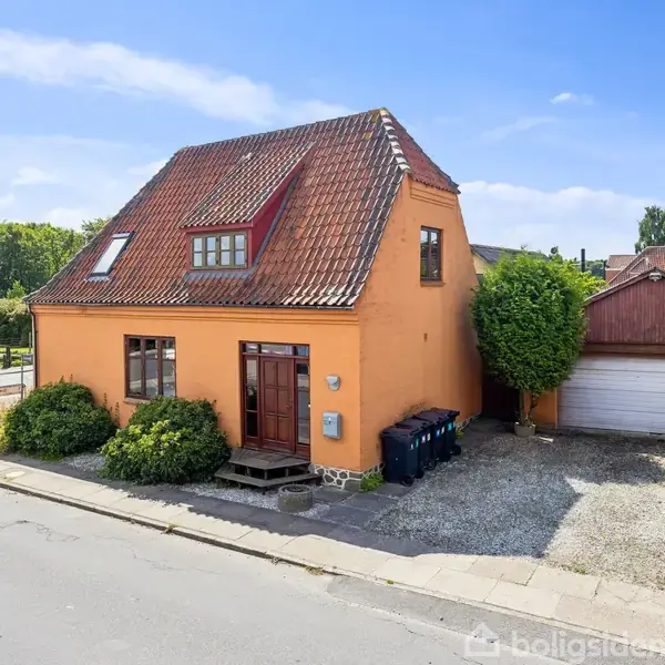 Et orange hus med rødt tag står ved en vej, omgivet af buske. Ved siden af huset er en grusindkørsel, der fører til en trægarage. Det er solskinsvejr.