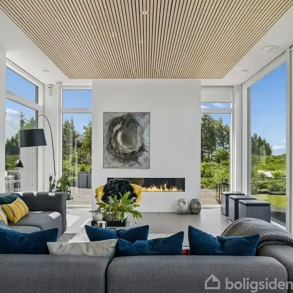 Stue med moderne sofaer og pejs. Store vinduer viser en skovrig, grøn udsigt. Kunstværker hænger på væggene. Elegant og stilfuldt interiør med naturligt lysindfald.