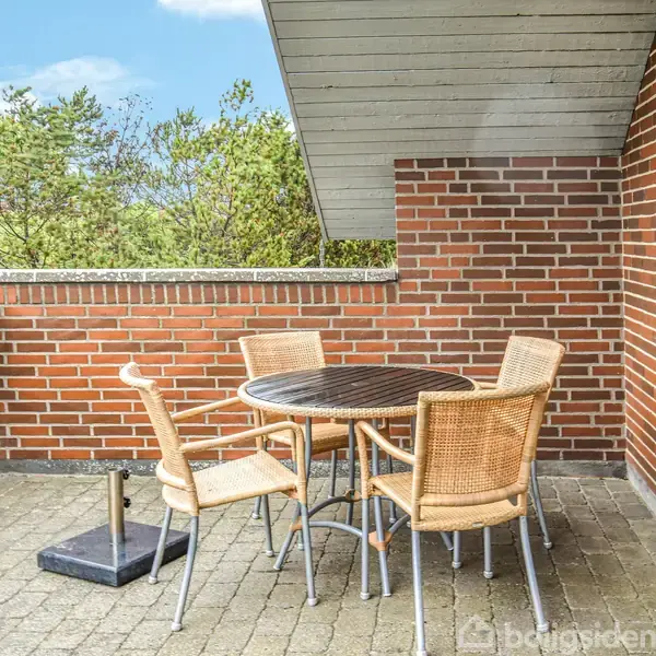 Rundt havebord med fire stole på en terrasse. Baggrunden viser røde murstensvægge, og omgivelserne er træer og en blå himmel.