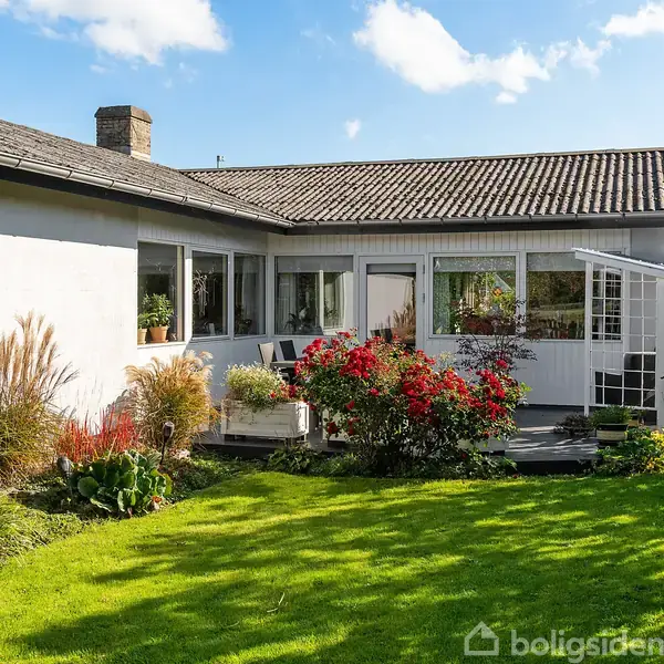 Hus med fladt tag og hvid facade står i solrig have. Røde og grønne planter omringer terrassen med havemøbler. Klart blå himmel med få skyer ses i baggrunden.