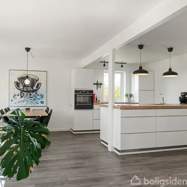 Et moderne køkken med hvide skabe og træbordplade, hvor to hængelamper lyser området op. I baggrunden er der et spisebord og en stor vægplakat. Grøn stor plante til venstre.