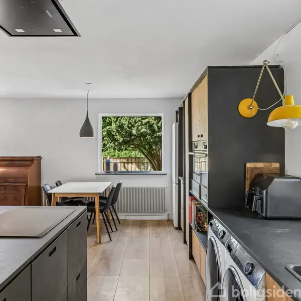 Køkkenet er elegant indrettet med sorte skabe og moderne apparater. En gul pendellampe hænger over bordpladen, mens et spisebord står ved vinduet, hvorfra der ses et træ udenfor.
