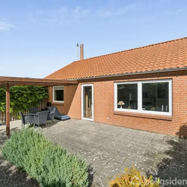 Rødt murstenshus med rødt tegltag står stationært. Foran huset er en flisebelagt terrasse med havemøbler. Haven er omgivet af planter, og der er en pergola på venstre side.