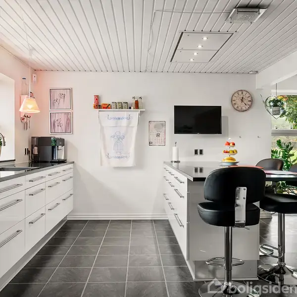 Køkken med lys bordplade, høje sorte stole ved en ø, røde lamper over bordpladen, og vægur på væggen. Vindue med udsigt til grønt.