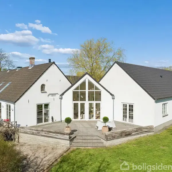 Et hvidt, moderne hus med store vinduer står i en frodig have. Der er en terrasse foran huset med stole og planter. Blåt himmel med spredte skyer ses ovenfor.