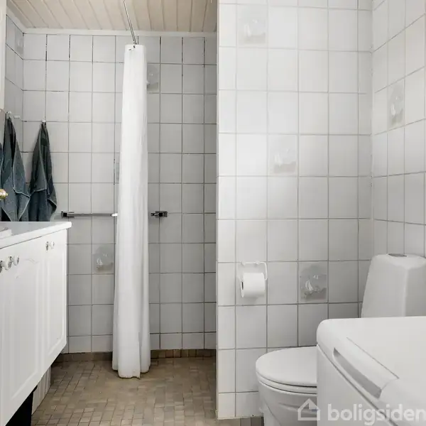 Et hvidt badeværelse med bruser bag forhæng, toilet og vask med skabsplads. Håndklæder hænger ved vasken. Rummet er flisebelagt. Dørskilt markerer "toilet" på en halvåben dør.