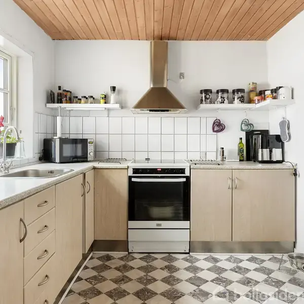 Et køkken med lyse træskabe langs væggene, en stålvask og mikrobølgeovn til venstre. En ovn med emhætte placeret i midten, omgivet af hvide fliser, og åbne hylder med krydderiglas.