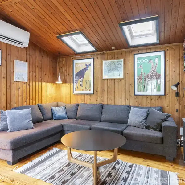 En grå hjørnesofa står i en træbeklædt stue med skråloft og loftvinduer. Væggene har plakater, og der er en reol med bøger. Et rundt sofabord og tæppe foran sofaen.