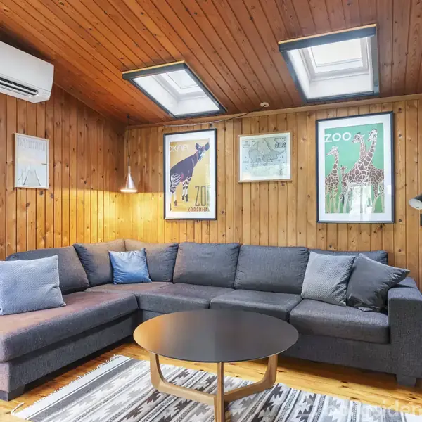 Stor grå hjørnesofa med puder står i en stue med trævægge og loft. Ovenlys, plakater, luftkonditionering, reol og et rundt sofabord præger rummet. Bøger og lamper ses omkring.
