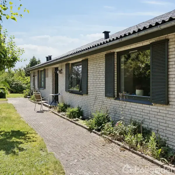 Et enfamiliehus med lyse mursten har to store vinduer med skodder. En stensti omgivet af græs og træer fører til en terrasse med stole, under en klar himmel.