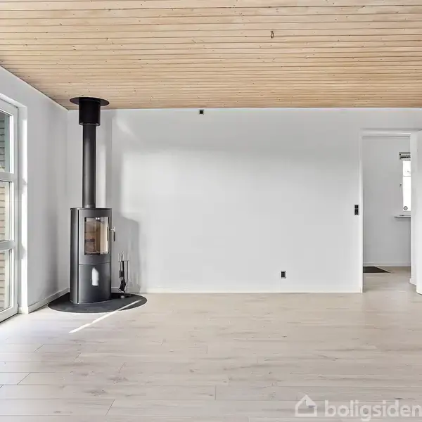 Brændeovn står stille på lys trægulv, omgivet af hvide vægge og vinduer med udsyn til have. Rummet har åbent loft i træ og en åben dør til venstre.