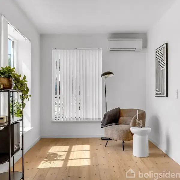 En stue har en beige lænestol ved et lille rundt bord, oplyst af et høj gulvlampe. Hylde med planter og nips står til venstre, og lys strømmer gennem vinduet.