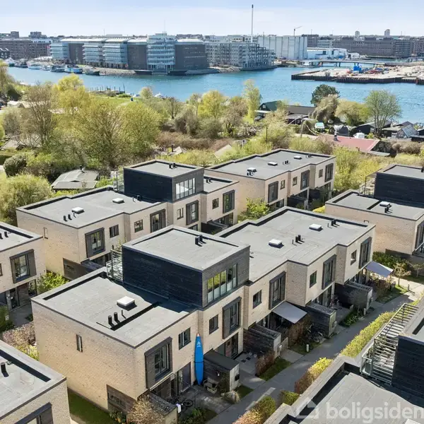 Moderne rækkehuse står i rækker med flade tage, omgivet af grønne områder. I baggrunden ses en blå bugt og en bylinje med industri- og boligbyggeri.