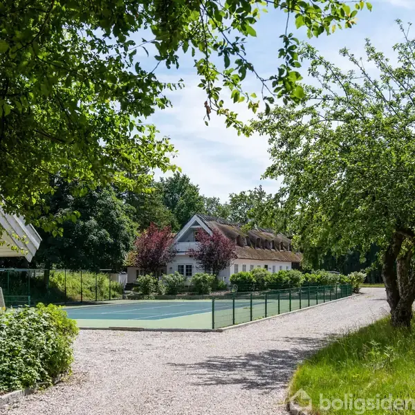 En grusvej fører til et tennisbane med et hvidt hus i baggrunden, omkranset af frodige træer og grønne buske under en lys himmel.