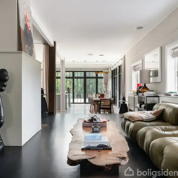 En sort skulptur står stille ved en hvid væg i en moderne stue med en grøn sofa og et træbord. Store vinduer bagtil åbner op til en grøn udsigt.