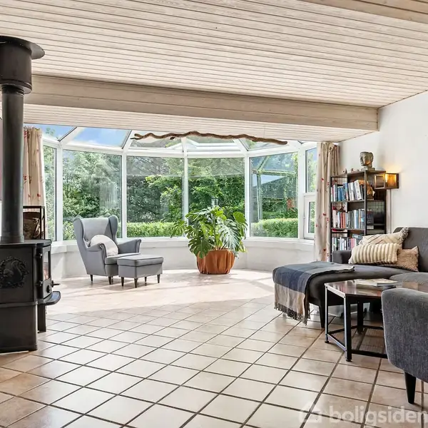 Stue med mørk brændeovn til venstre, stor grå sofa med puder til højre. Glasvæg bagved afslører terrasse og have. Flisegulv og træloft understreger den lyse og rummelige atmosfære.