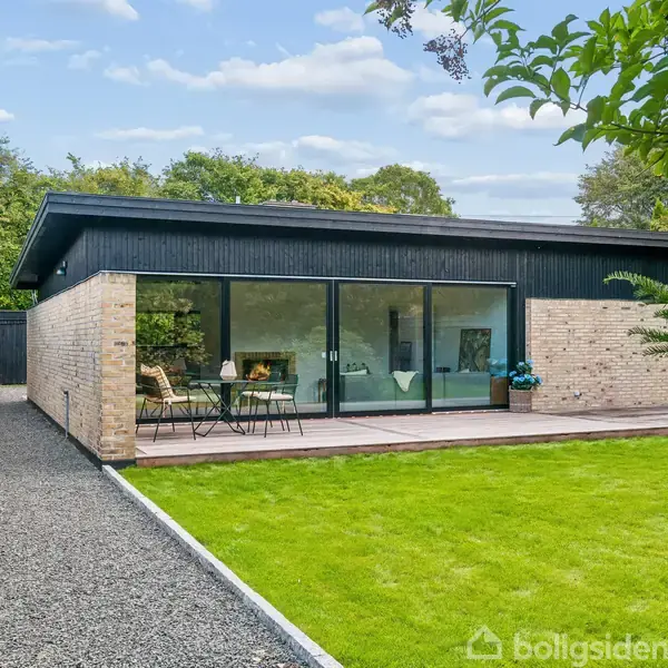 En moderne bungalow med store vinduer står i en grøn have. Foran er der en terrasse med havebord og stole. Grussti fører op til huset. Ingen tekst i billedet.