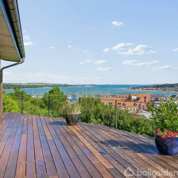 Træterrasse med udsigt over en by og en havnefront. To plantede krukker står på terrassen. Solrigt vejr med få skyer på himlen.