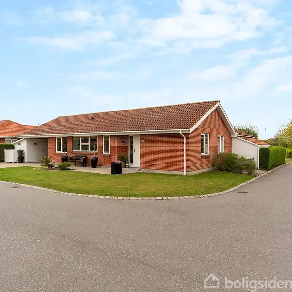 En rød murstensvilla med rødt tegltag, placeret på et hjørne i et roligt boligområde. Villaen omgives af en frodig have med trimmede buske under en blå himmel.