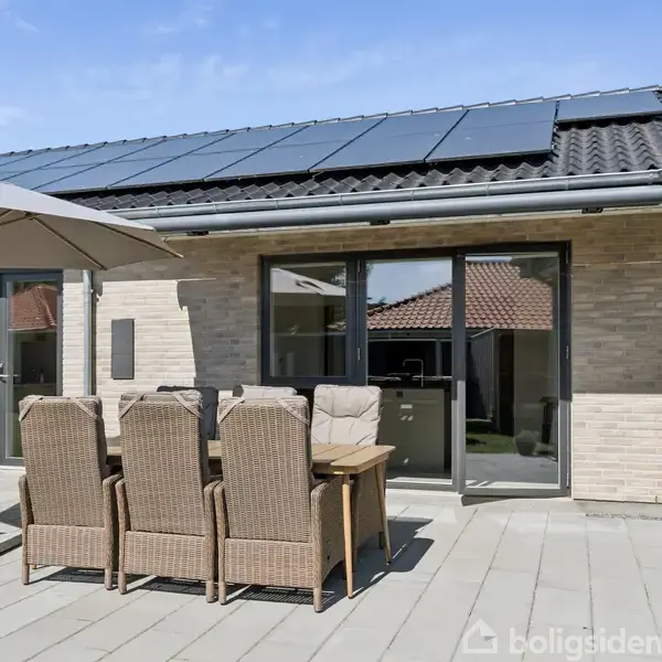 En udendørs terrasse med seks rattan-stole og et bord står under en stor parasol. Solcellepaneler er installeret på taget af et moderne hus med store vinduer.