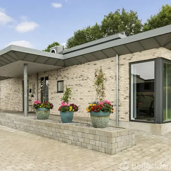 Et moderne hus med fladt tag og lyse mursten, omgivet af blomsterkrukker på en flisebelagt indkørsel. Store vinduer giver indblik i rummene, mens grønne træer danner baggrunden.