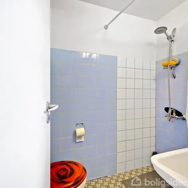 Et badeværelse med en bruser i et hjørne. Væggene har lyseblå fliser. En badeforhæng dækker bruseområdet. Toilettet har et sæde med en rød blomstret pude.