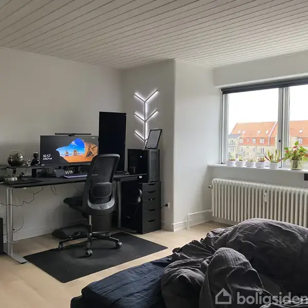 En kontorstol står ved et stort skrivebord med computerudstyr, mens dagslys strømmer ind gennem et vindue. Vækkeuret viser "17:00". Indendørs plante og minimalistisk indretning præger rummet.