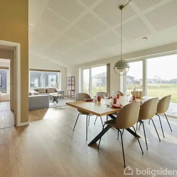 Spisebord med seks stole står i et lyst rum med store vinduespartier, der viser en græsplæne og bygninger udenfor. En sofa og en åbent dør til soveværelse ses i baggrunden.