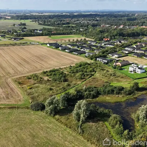 Et åbent, landligt område med en stor mark ved siden af en lille sø. I baggrunden ses en landsby med boliger og skovområder spredt rundt i landskabet.