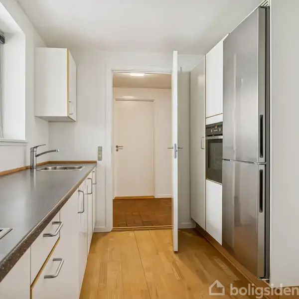 Køkken med indbyggede hvidevarer og skabsplads langs væggen, træbordplade og vindue på venstre side, træparketgulv, dør til anden rum for enden.