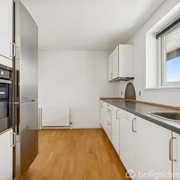 Et moderne køkken har hvide skabe, en sort bordplade og en indbygget ovn ved siden af et køleskab. Et vindue giver udsigt og lyser rummet med trægulve op.