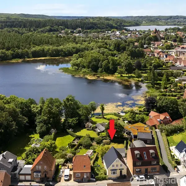 Hus med gråt tag og rød murstensfacade markeres med en rød pil. Det står blandt andre huse tæt på sø og grøn skov i en by.
