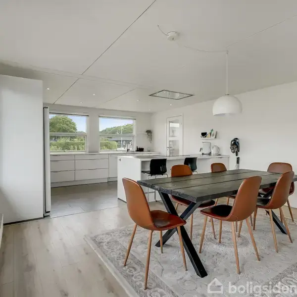 Et moderne spisebord med brune stole står på et tæppe i en lys spisestue. Baggrunden viser et åbent køkken med hvide skabe og store vinduer med udsigt til grønne træer.