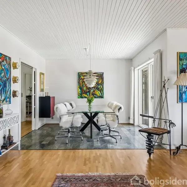 Et spisestuebord omgivet af fire stole står på en mørk flisebelægning. Omgivet af abstrakte malerier og en stående lampe, indrammer en glastdør omgivelserne i det lyse rum.