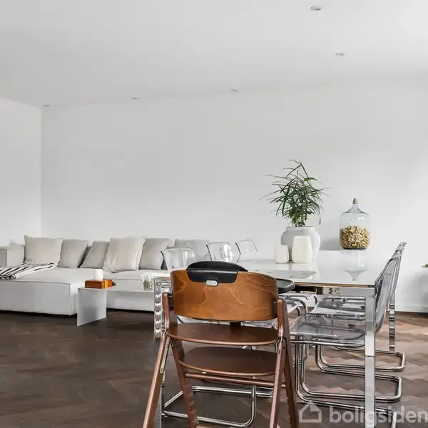 En moderne stue med en hvid sofa fyldt med puder står i baggrunden. Foran er et spisebord med en stor glasbeholder og planter. Lyse trægulve og hvidmalede vægge.
