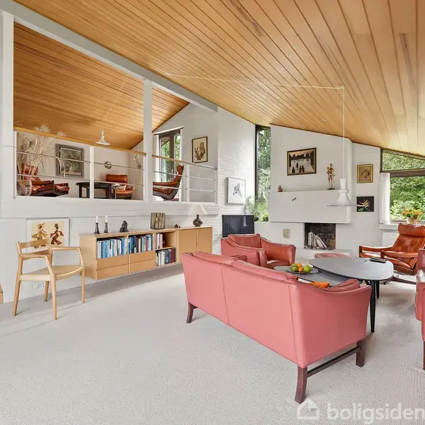 Stue med røde sofaer og sofabord, placeret foran en pejs. Træloft og trapper fører til forhøjet område med reoler og maleri. Store vinduer med udsigt til natur.