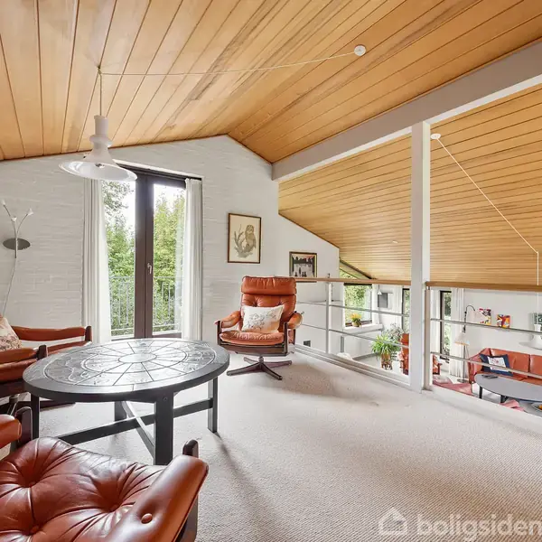 Et loftsrum med træloft og tæppebelagt gulv rummer to lænestole og et rundt bord. Vinduerne giver udsigt til frodig have. Trappeadgang til stuen nedenfor.