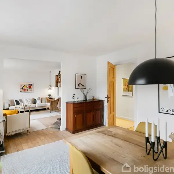 Et spisebord med stearinlys står i forgrunden; en sort lampe hænger over. I baggrunden ses en stue med møbler, planter og kunstværker på væggene, som skaber en hjemlig atmosfære.