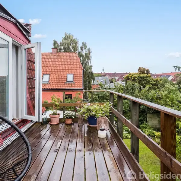 En træterrasse rummer plantede potter på rækken; åben fransk dør åbner mod rød tagstensbygning. Grønne buske og træer pryder baggrunden under en blå himmel.