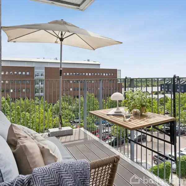 En balkon har en sofa med puder, et træbord med en lampe og planter, mens en parasol skygger. Udsigt til en bygning og parkeringsplads i et byområde.