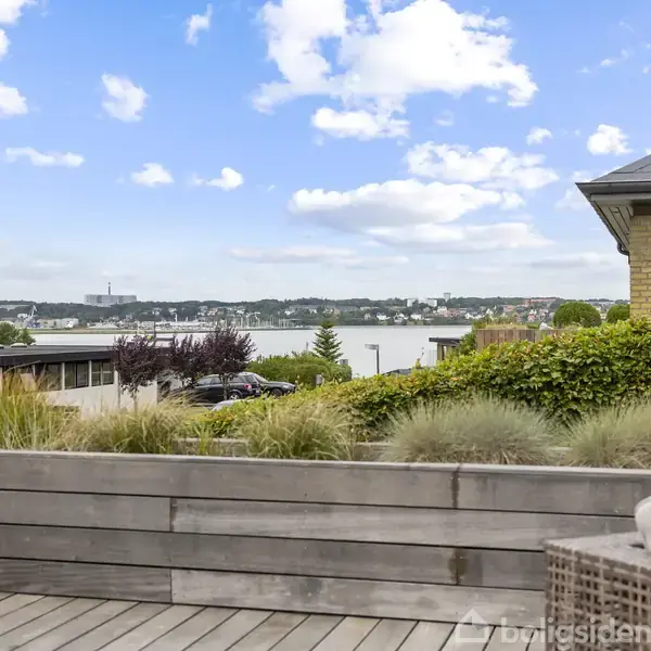 Terrasse udført i træ med sofa og udsigt over en sø og by i baggrunden, omgivet af grønne buske og en blå skyet himmel.