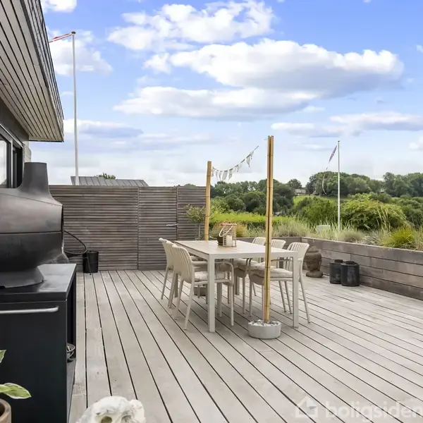 En moderne terrasse med trædæk har et spisebord og stole centreret omkring det. En pejs er placeret på en skænk. Omkring er der grøn beplantning og klare, blå himmel.