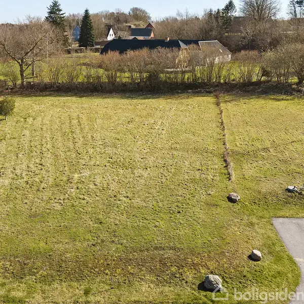 Græseng ligger stille, omkranset af buskadser, med en lille vej i forgrunden. I baggrunden står et par huse blandt træer, omgivet af åbne marker.