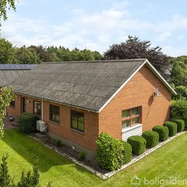 Et rødt murstenshus med skråt tag og solpaneler. Det står i en velplejet have med grønne buske og græs. Omgivet af træer under en klar blå himmel.