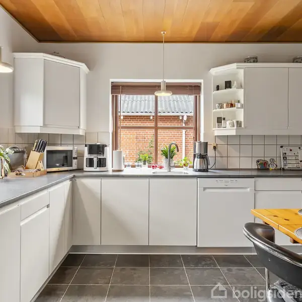 Køkken med hvide skabe og gråt gulv har apparater og planter på bordpladen. Et stort vindue bagved giver udsigt til en murstensbygning. Runde dækkeservietter på et træbord.