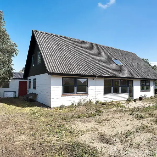 Hvidt hus med sort tag står på en tør græsplæne. Huset har store vinduer og et skråt tag med ovenlysvindue. Træer og hække omkranser ejendommen under en blå himmel.