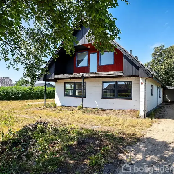 Et hvidt hus med røde vinduer står solbelyst i en have med en grusindkørsel. En carport er tilknyttet, omkranset af hække. Skyfri blå himmel ses i baggrunden.