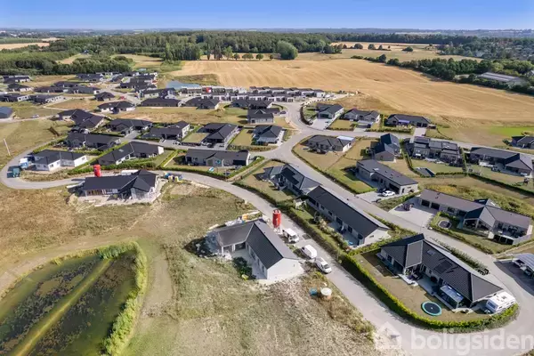 Boligkvarteret viser huse med sorte tage placeret langs snoede veje. Omkringliggende marker og skove danner en naturskøn baggrund. Nogle byggeprojekter er igang, og en lille sø er synlig.